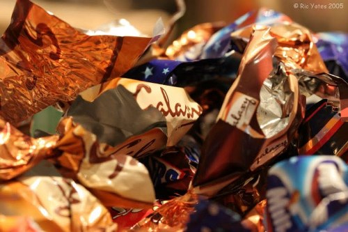 A close-up image showing a collection of crumpled foil wrappers and paper packaging in various metallic and matte finishes, predominantly in shades of copper, silver, and blue. The wrappers are piled together, creating a textured, chaotic arrangement with visible creases and folds. The background is blurred, indicating that the focus is on the discarded packaging materials, likely on a flat surface such as a table or pavement. This scene illustrates waste items typically handled during a rubbish removal service by House Clearance Berkhamsted, set in a residential or outdoor environment within or near Berkhamsted. The metallic sheen of the wrappers contrasts with the duller, flattened paper parts, emphasizing the mixed nature of household rubbish often collected for house clearance or rubbish disposal in the local area. The image’s natural lighting highlights the reflective surfaces and textured details of the packaging materials, aligning with a professional waste management context in Hertfordshire.