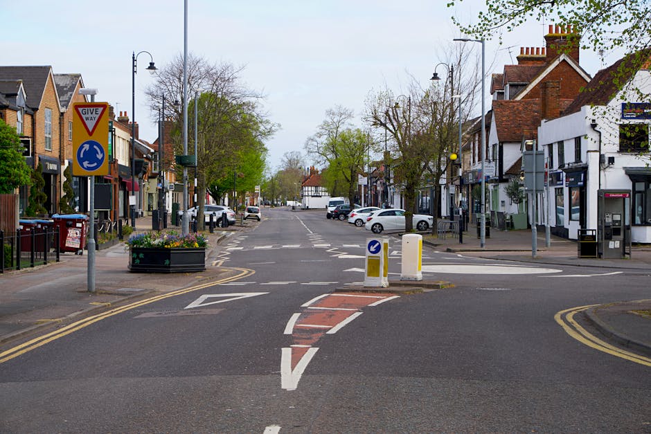A quiet street scene in a residential and commercial area, featuring a T-junction with traffic calming measures such as small white and yellow bollards and curved road markings indicating a change in